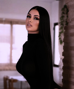 A long-haired brunette in a black bodysuit poses against a stone wall.