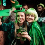 Two girls in a wig and hat make selfi at the bar. They celebrate St. Patrick's Day. They are having fun.