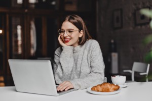 Cheerful girl in gray outfit working in laptop during lunch with croissant and cup of coffee