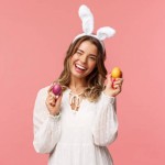 Holidays, spring and party concept. Cheerful good-looking blond woman celebrating Easter day in rabbit ears, holding two painted eggs and wink camera, smiling happily, pink background.