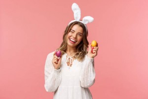 Holidays, spring and party concept. Cheerful good-looking blond woman celebrating Easter day in rabbit ears, holding two painted eggs and wink camera, smiling happily, pink background.