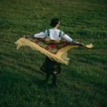 Back view of a woman in an embroidered shirt holding a bright floral shawl outdoors.
