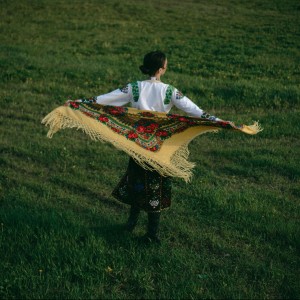 Back view of a woman in an embroidered shirt holding a bright floral shawl outdoors.