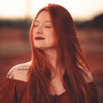 Red-haired woman with eyes closed wearing a burgundy off-the-shoulder top outdoors.