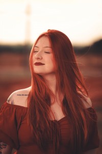Red-haired woman with eyes closed wearing a burgundy off-the-shoulder top outdoors.